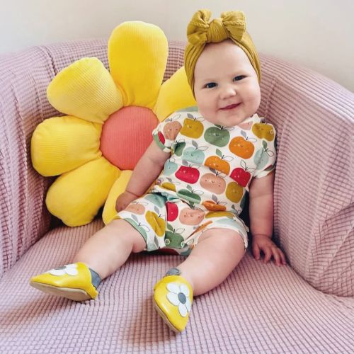 Toddler girl wearing yellow Dotty Fish barefoot shoes with grey ankle trim and white flower design, sitting with flower cushion.