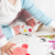 Toddler reading a book, wearing white leather Dotty Fish first walker shoes with pink ankle trim and multicoloured spots.