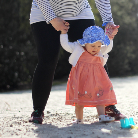 Toddler taking first steps on sandy beach, wearing her white flower sandals.